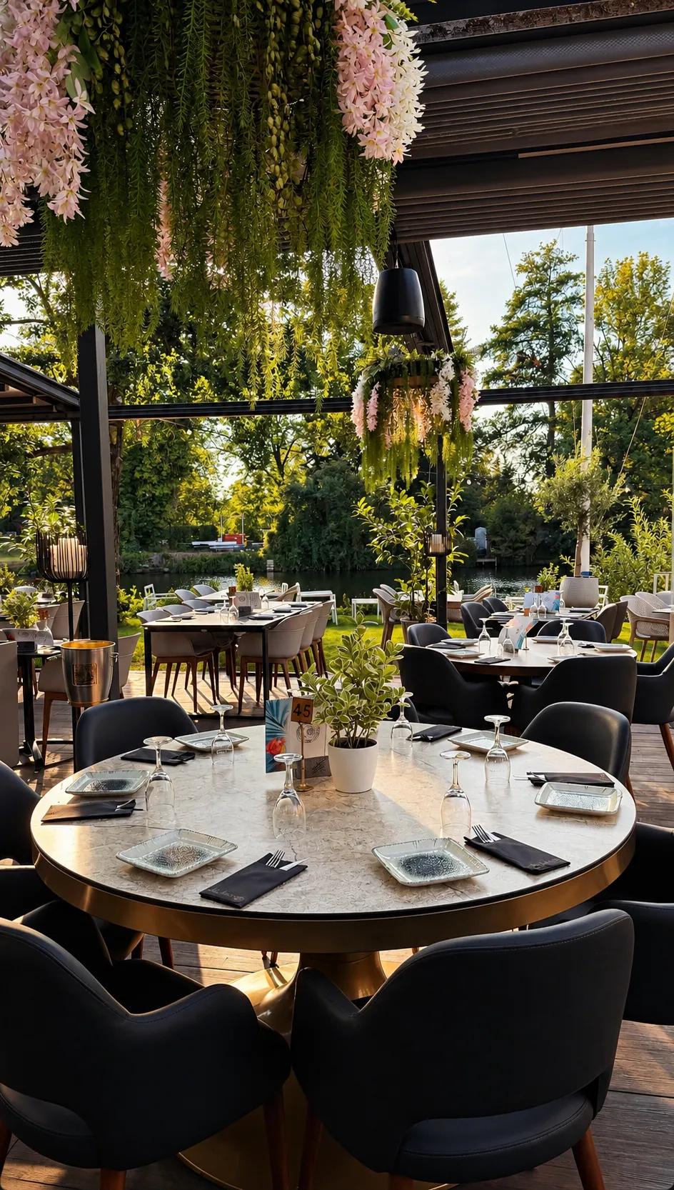 Gedeckter runder Tisch auf der Terrasse des Âu Lac unter der Pergola mit Blütenranken, dahinter Bäume und der Fluss im Abendlicht